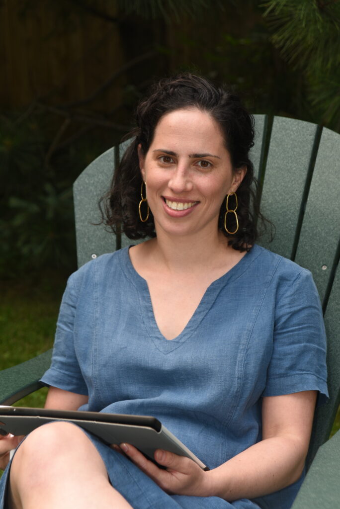 Photo of Catherine Jampel. White woman in late 30s with shoulder-length brown hair, brass earrings, casual blue dress, sitting on dark green Adirondack-style chair and holding a notepad of some kind.